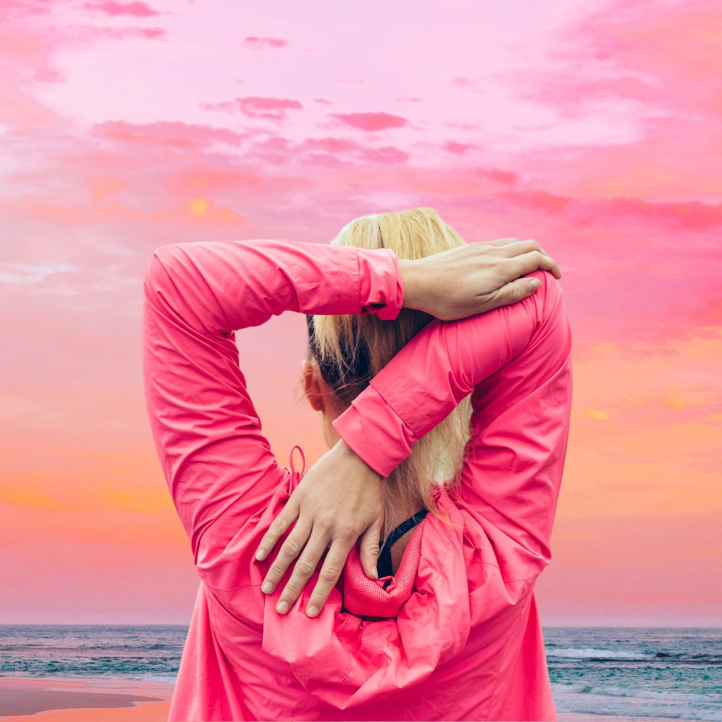 photo d'une femme, au bord de la mer, vue de dos qui étire son coude en arrière de sa tête avec son autre main pour signifier la flexibilité et la mobilité retrouvée grâce aux thérapies manuelles